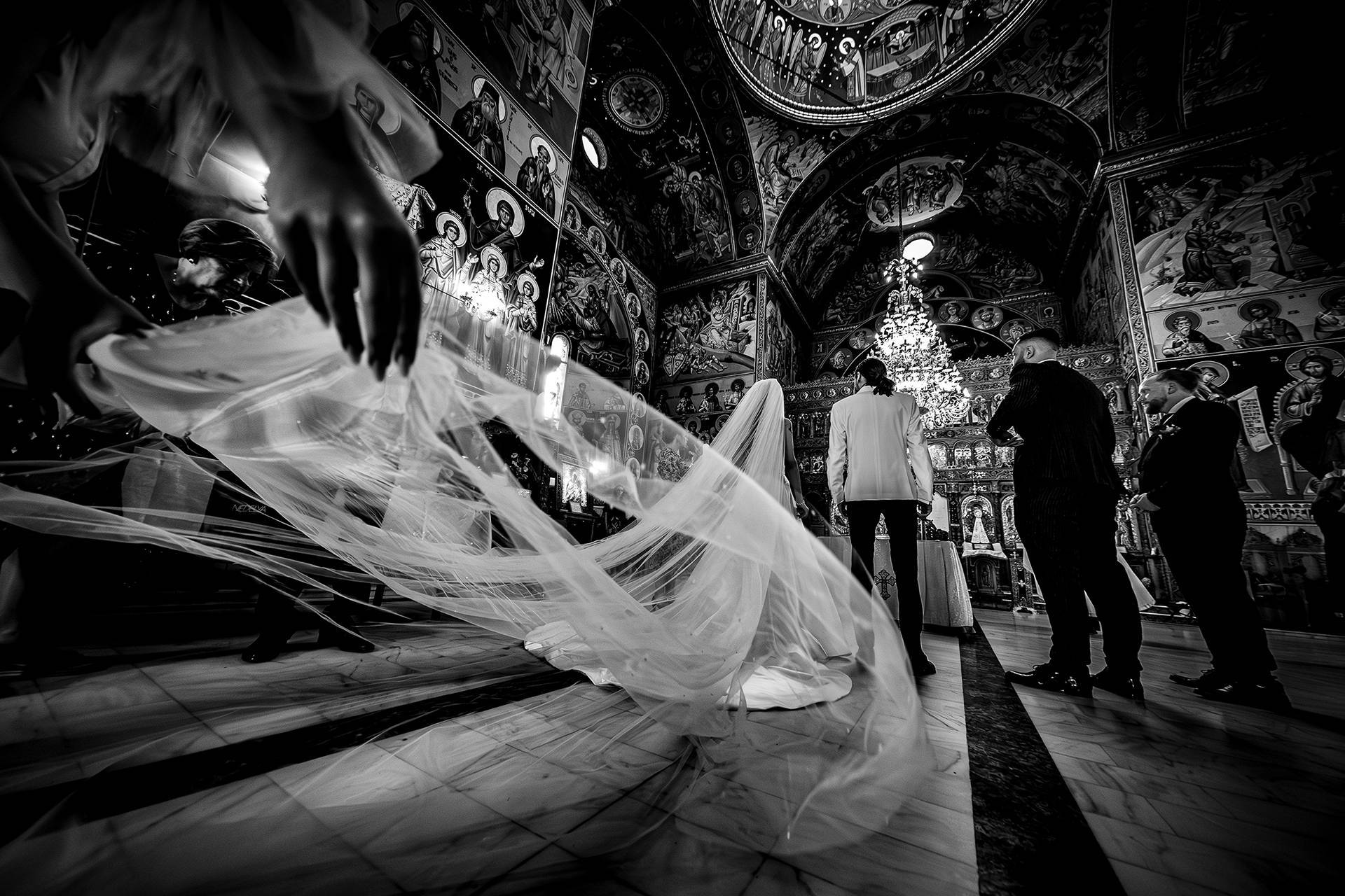 Voalul purtat cu eleganță – fotograf nuntă Transilvania Mirii cu spatele în biserică, iar voalul miresei este purtat de domnișoara de onoare – fotograf nuntă Transilvania