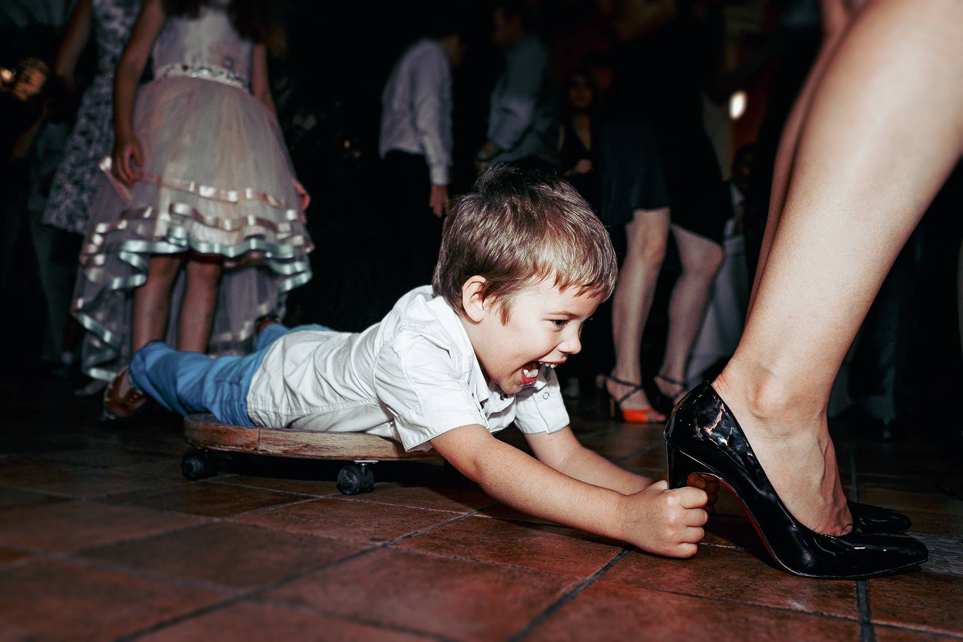 Joacă și energie la nuntă – fotograf nuntă Transilvania Copil se joacă pe o planșă cu roți, ținându-se de tocul unei femei care dansează la nuntă